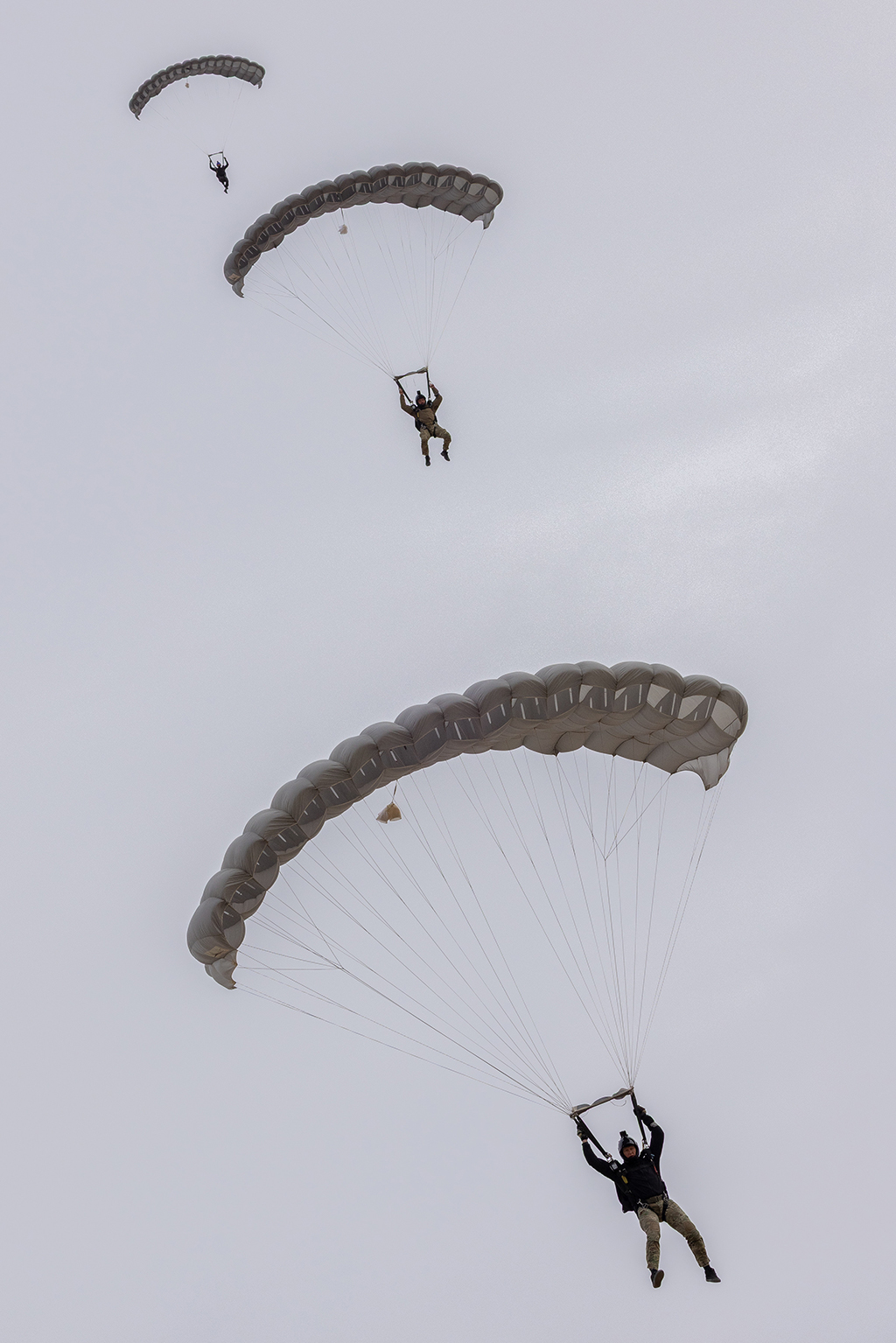 Three of the six Exercise Rising Gains 1‑26 parashutists continuing to descend with open grey coloured rectangular parachutes, closer to the ground in line astern.
